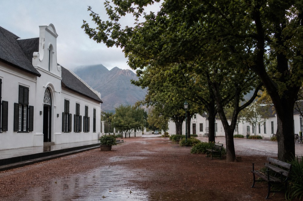 A compact town square with historic whitewashed buildings, benches under oak trees and mountains behind in soft afternoon light, an empty pavement, no signage, no text, no people
