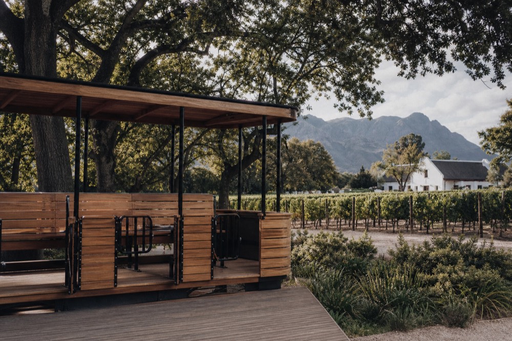 A wooden tram platform beside vineyards with oak trees and a gabled farm building in the distance, mountains behind, soft light, no people or signs