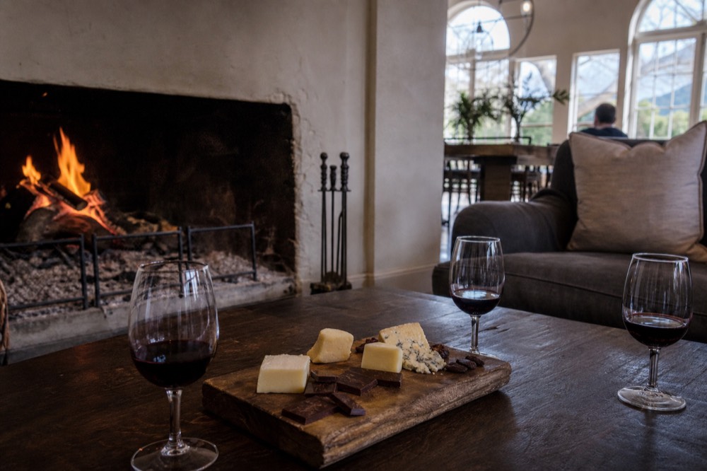 A cosy winter tasting room with a lit fireplace, a board of chocolate and cheese and glasses of red wine on a low table, warm light, no people, no text, no signage