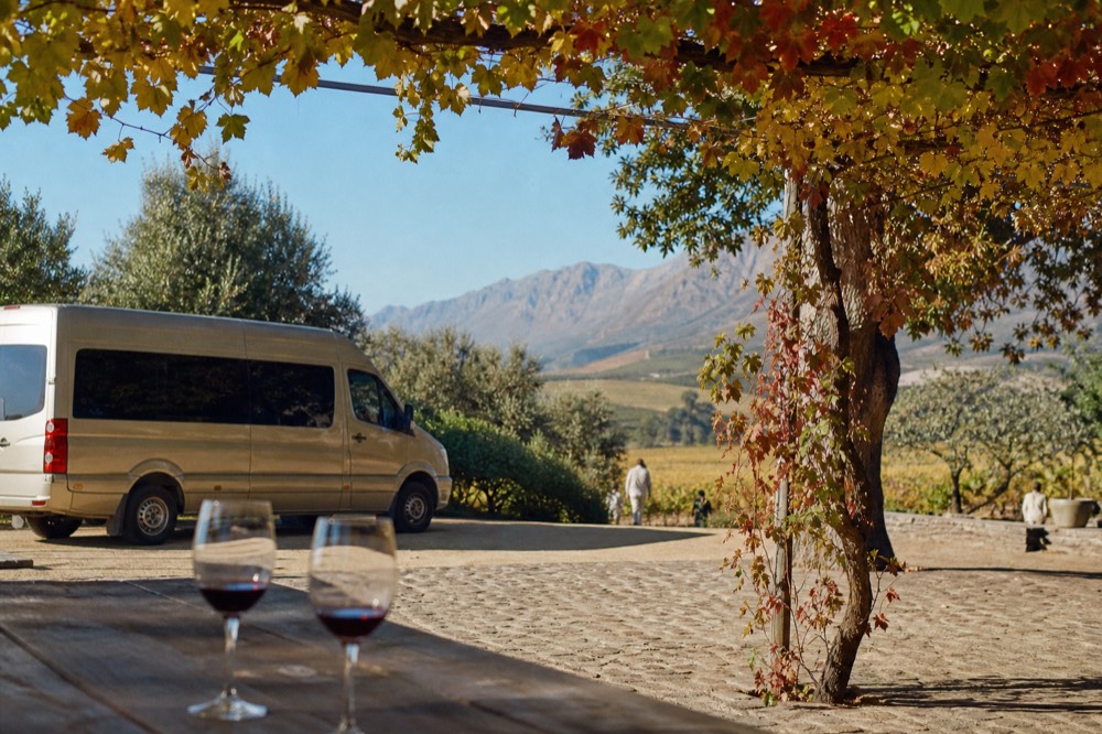 A small touring minibus parked beside a cellar-door courtyard shaded by vines, two wine glasses on a wooden table, mountains beyond, no faces or signage