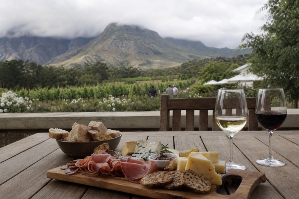 A spread of cheeses, charcuterie and bread on a wooden board beside two glasses of wine on an estate table, blurred green vineyard behind, no text, no logos, no faces