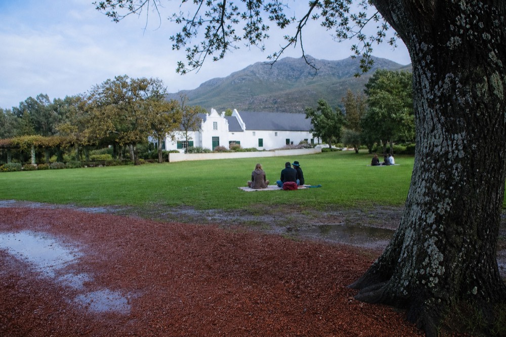 A broad green estate lawn with scattered oak trees and picnic blankets, a whitewashed farm building in the background under a blue sky, no branding, no text, no recognizable faces
