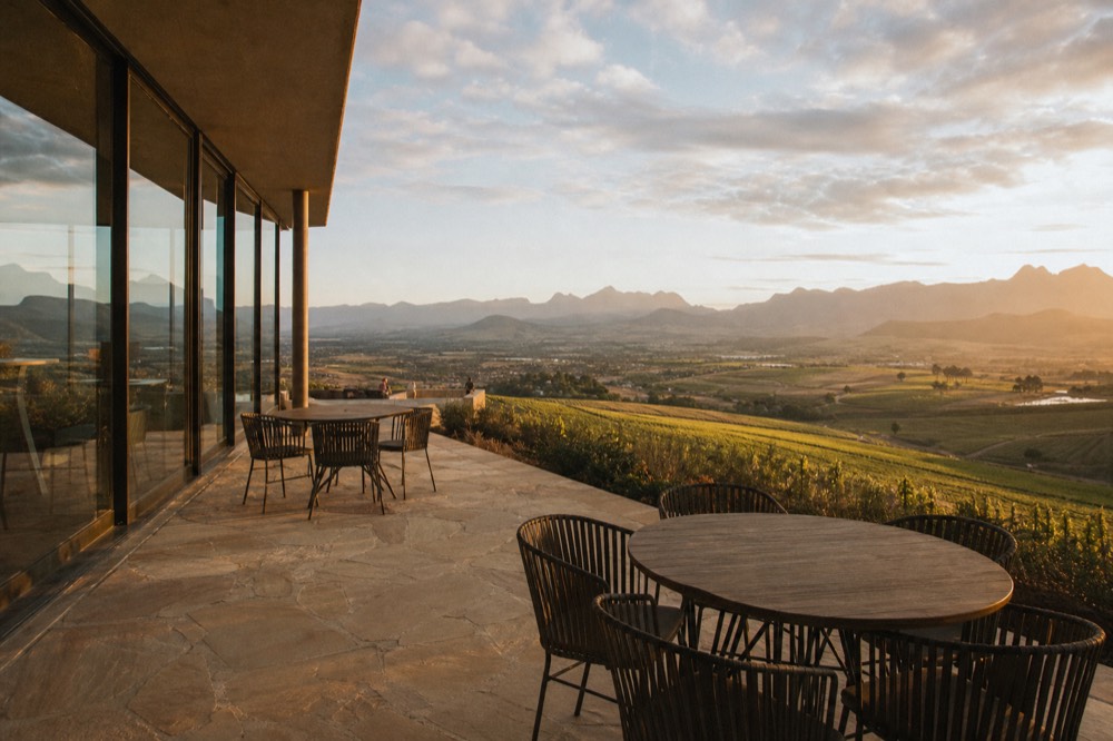 A glass-walled tasting room on a hilltop estate looking out over terraced vineyards and a distant valley at golden hour, empty seats on a stone terrace, no branding, no text, no people