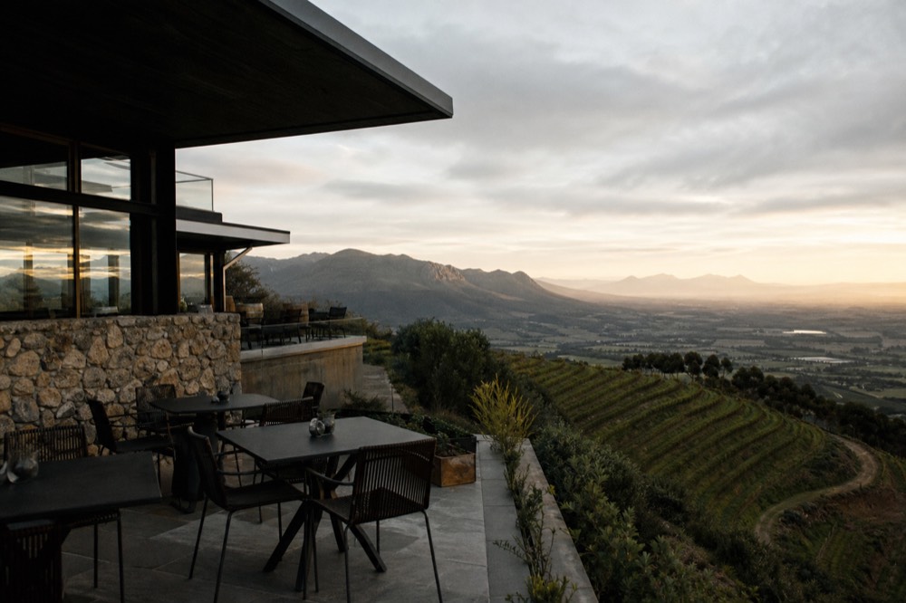 A modern glass-and-stone tasting terrace at the top of a mountain pass overlooking terraced vineyards and a distant valley at golden hour, empty chairs, no branding, no text, no people